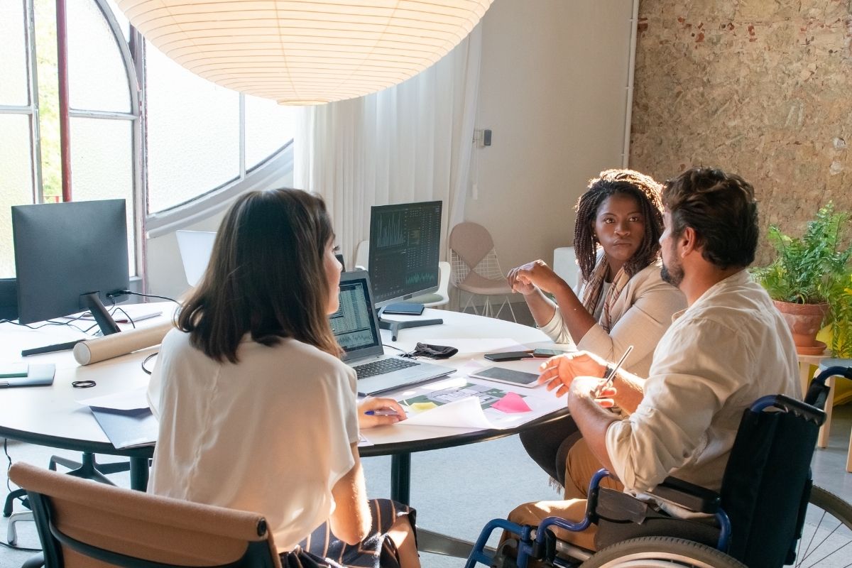 Team meeting in an office with a man using a wheelchair and two colleagues discussing documents and laptops around a table