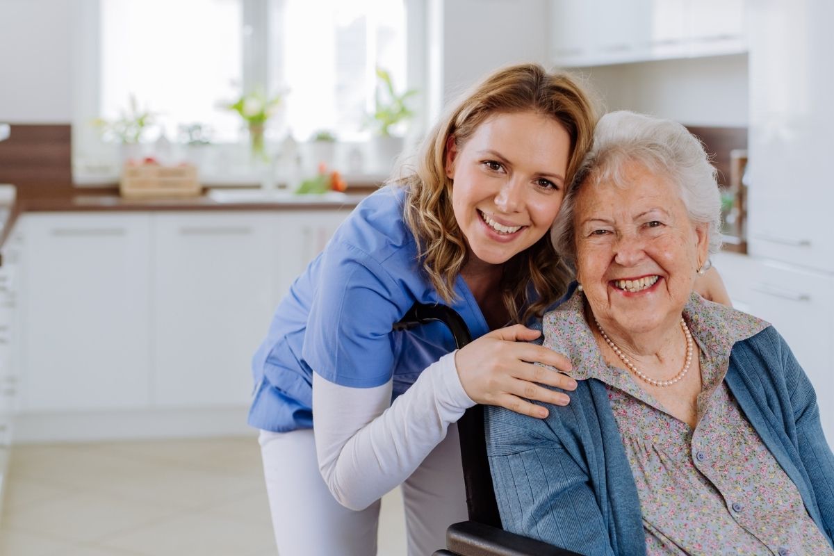 Smiling support worker with an older woman at home, both facing the camera in a kitchen