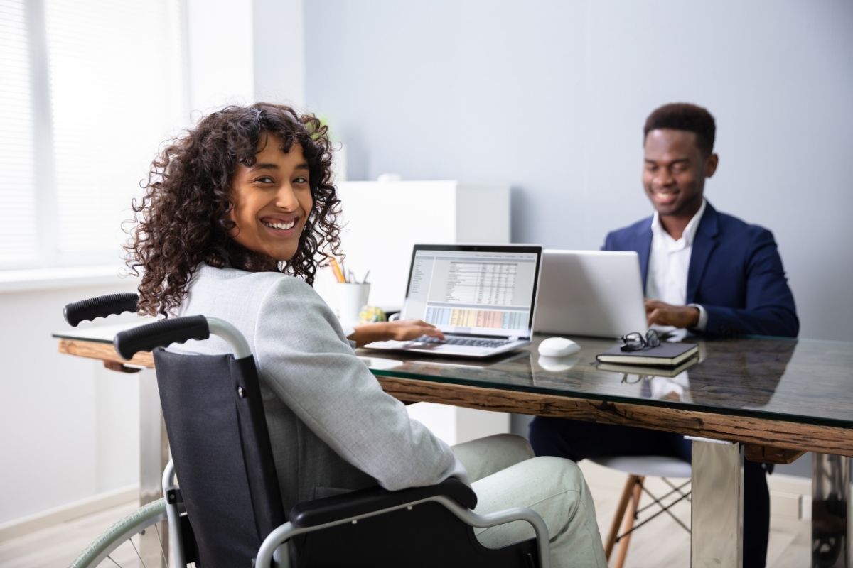 Professional woman using a wheelchair smiles at the camera while working on a laptop beside a colleague in an office