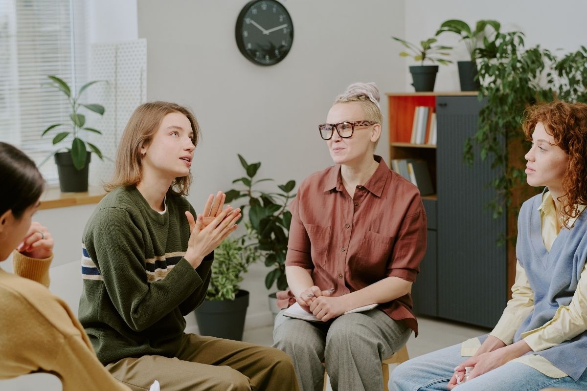 Adults in a small group therapy session, seated in a circle; one person speaks while others listen