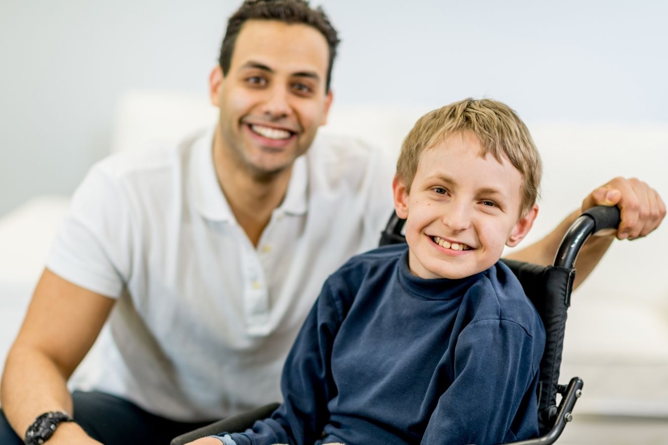 Smiling adult beside a young boy using a wheelchair at home