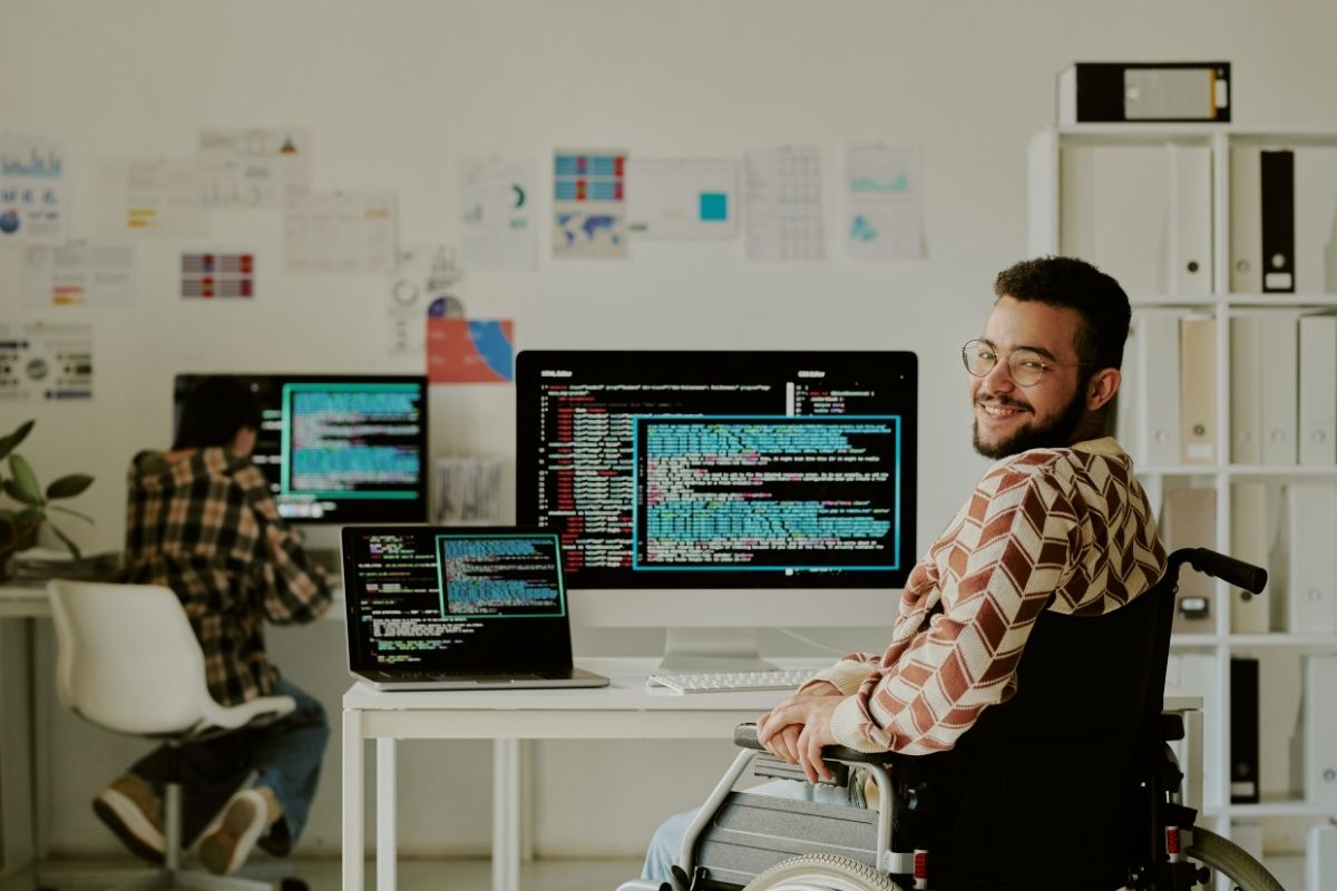 Developer using a wheelchair smiles at the camera in an office with multiple monitors showing code