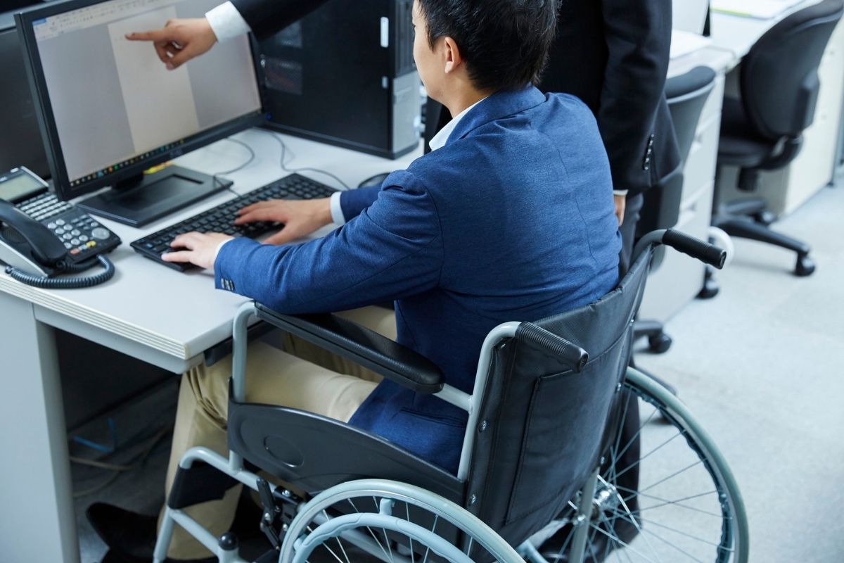 Office worker using a wheelchair typing at a desktop while a colleague points at the monitor