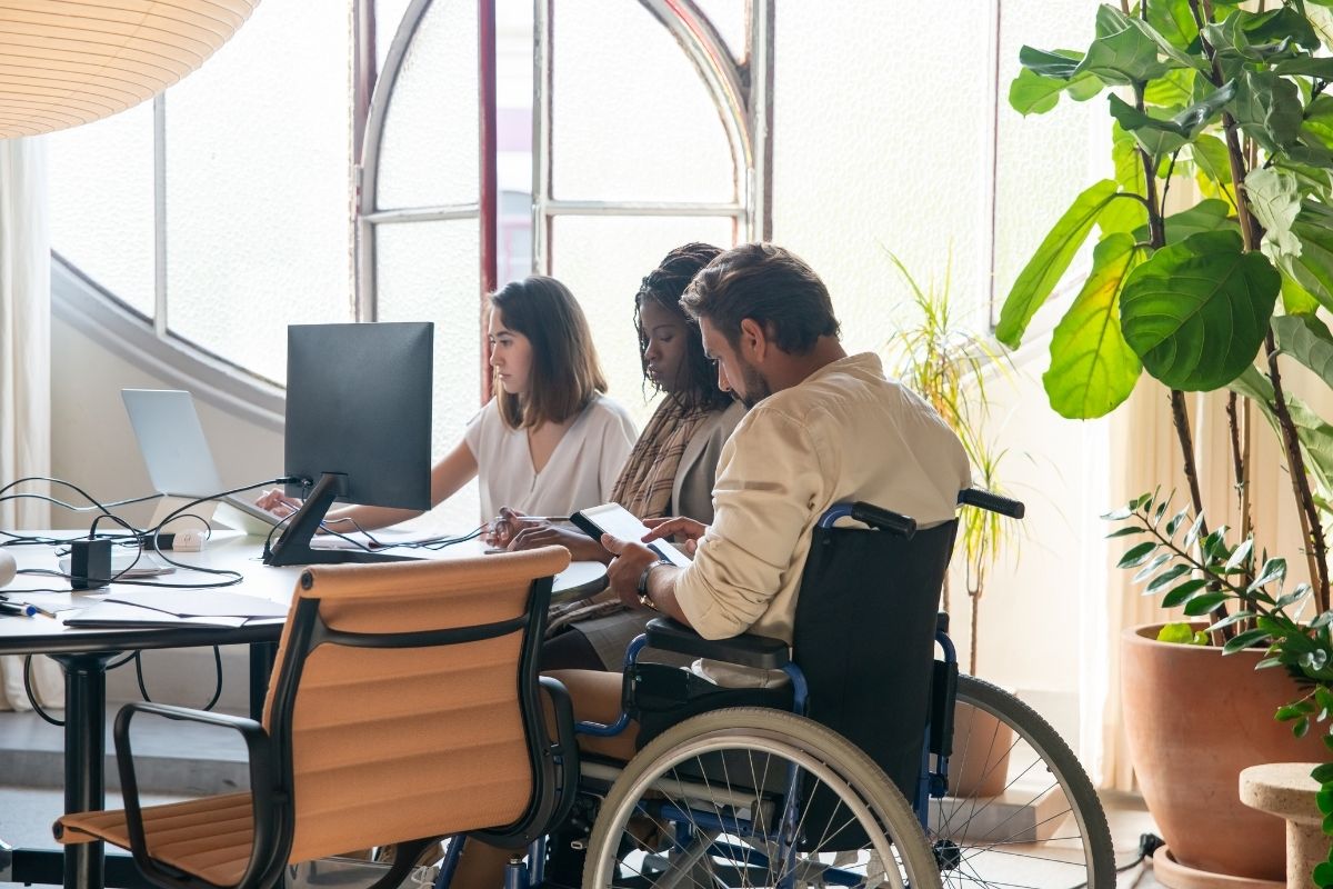 Team working at a desk in a bright office; a man using a wheelchair reviews documents with two colleagues
