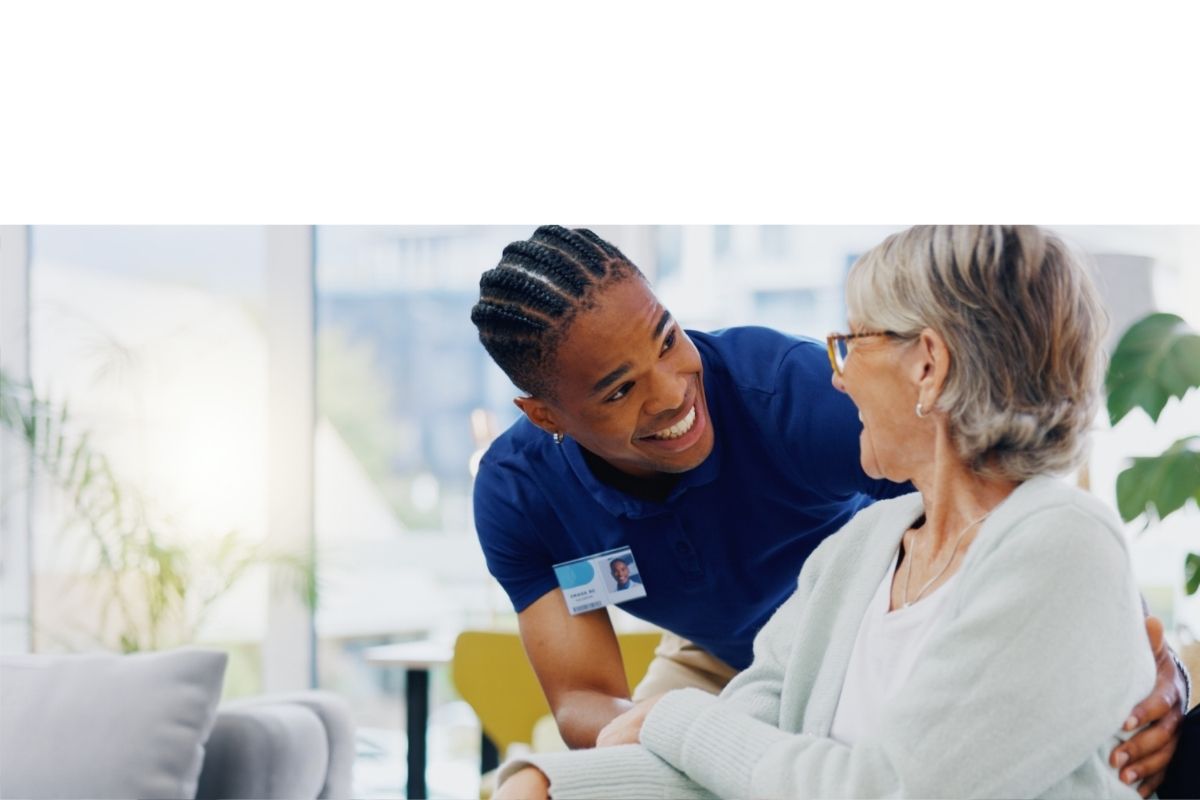 Support worker smiling and talking with an older woman in a wheelchair, an open book on her lap in a bright living room