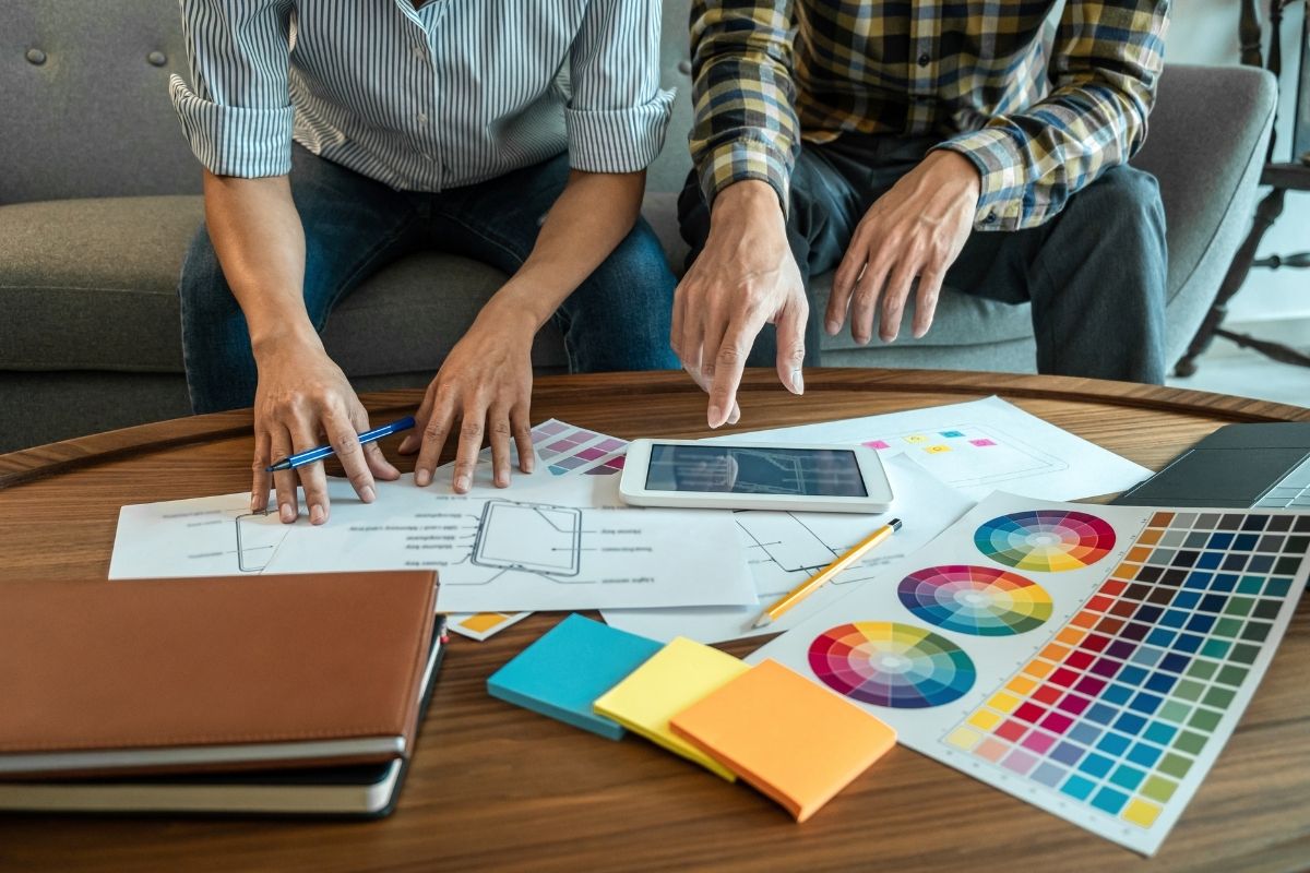 Two designers reviewing wireframes on a tablet with colour swatches and notes spread across a table