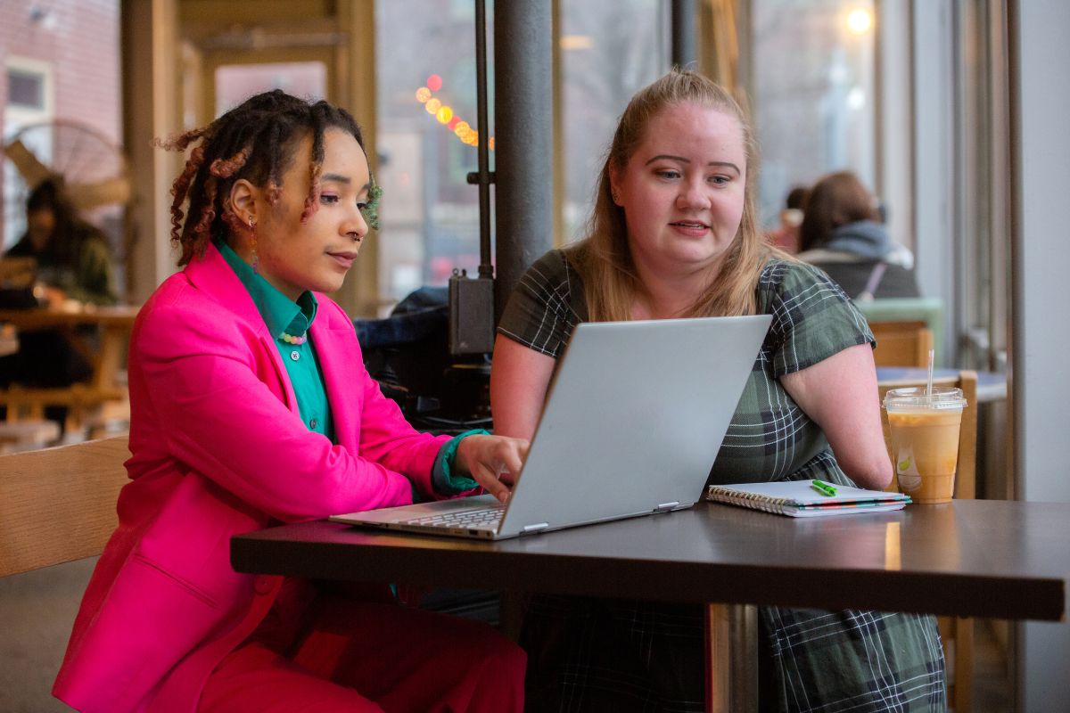 Two women collaborating on a laptop in a café, with notebooks and iced coffee on the table