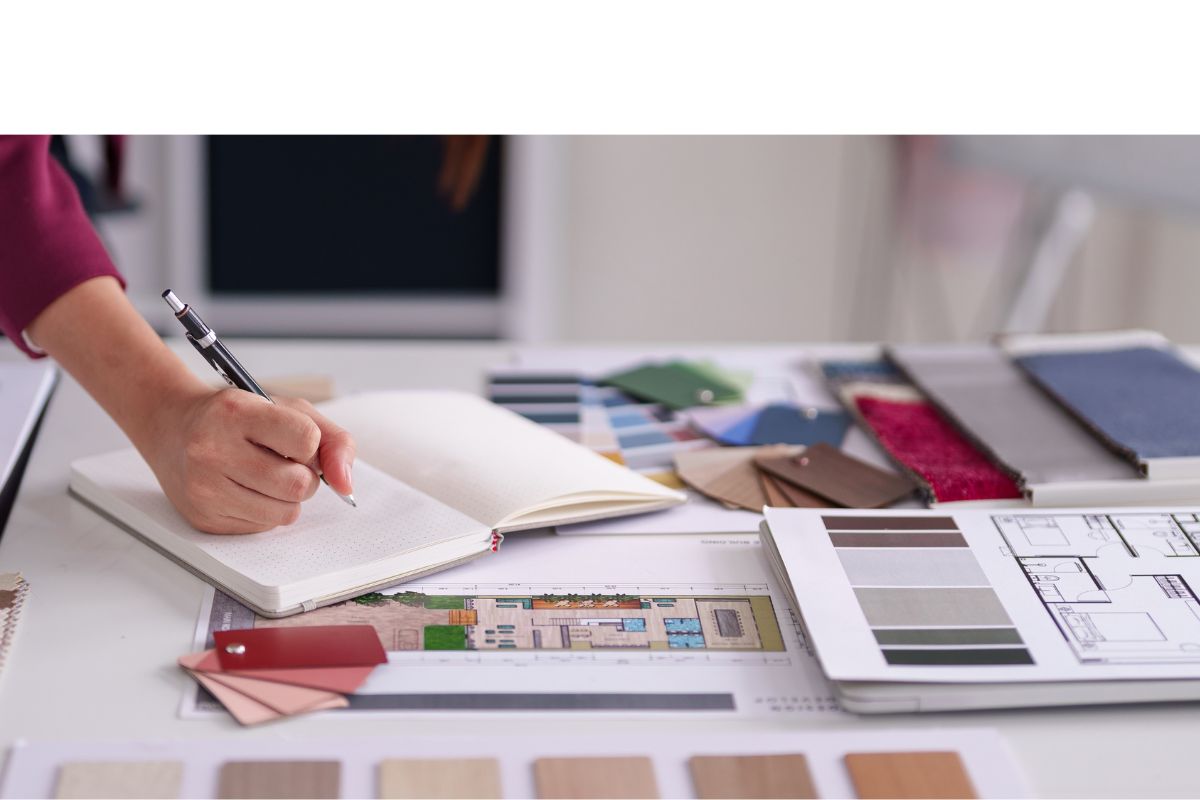 Interior designer writing in a notebook beside floor plans, colour swatches and fabric samples on a desk