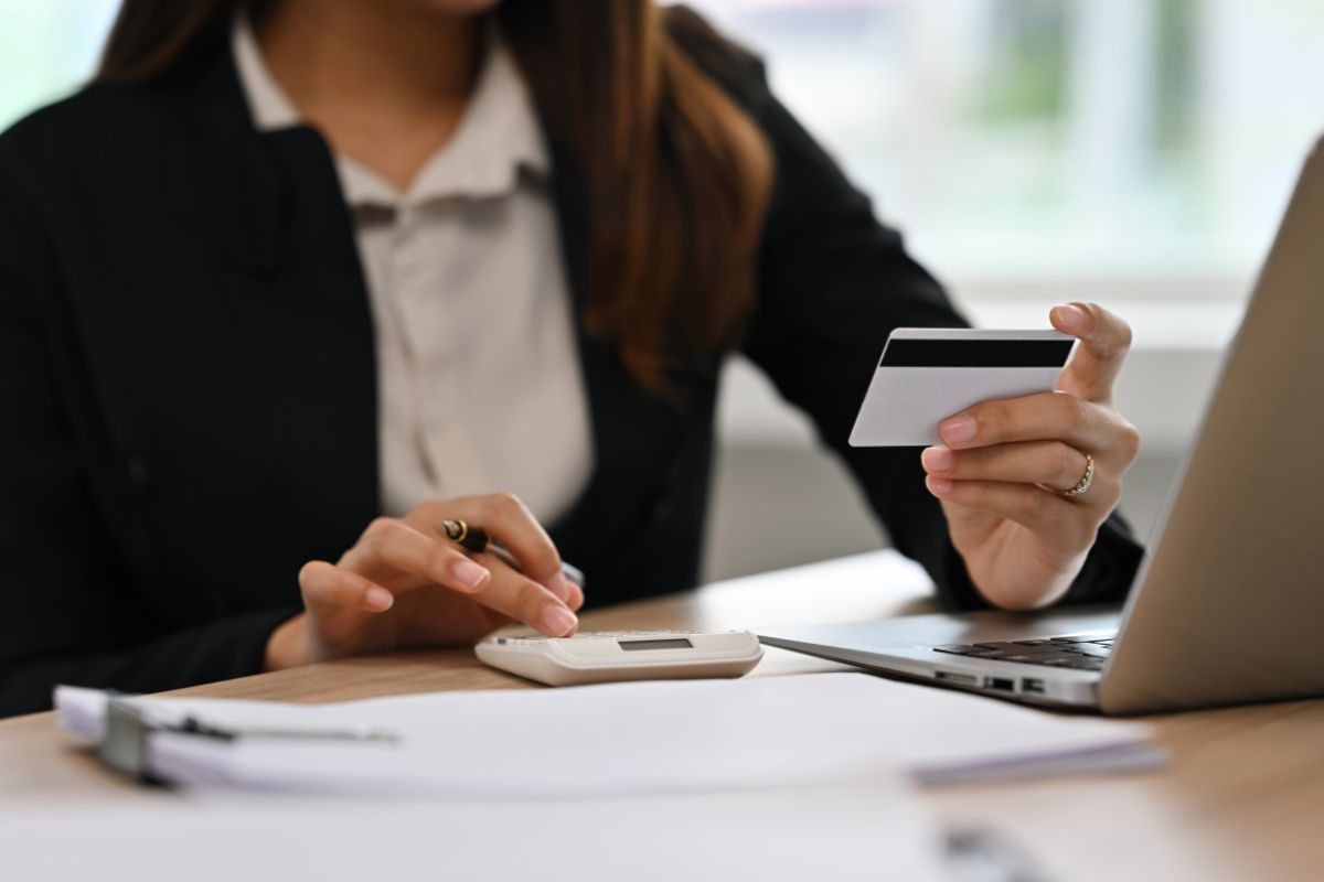 Woman holding a credit card and using a calculator beside a laptop at a desk