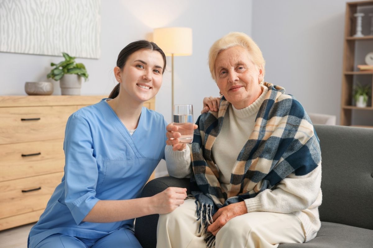 Support worker offering a glass of water to an older woman sitting on a sofa at home