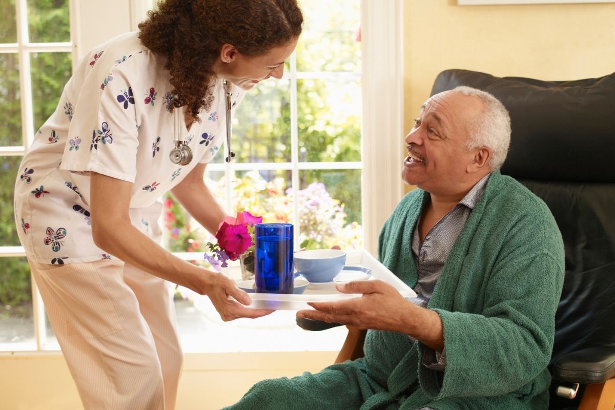 Care worker handing a tray with a drink and meal to an older man in a robe at home