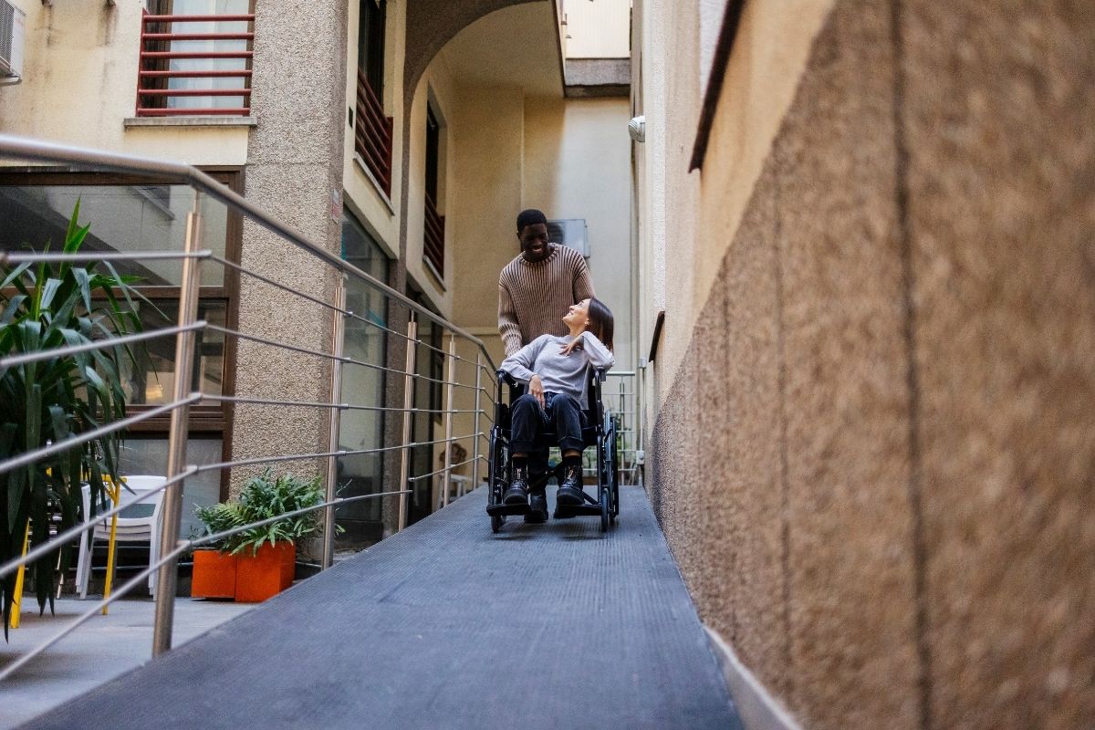 Support worker pushing an older adult in a wheelchair down an accessibility ramp in a building corridor