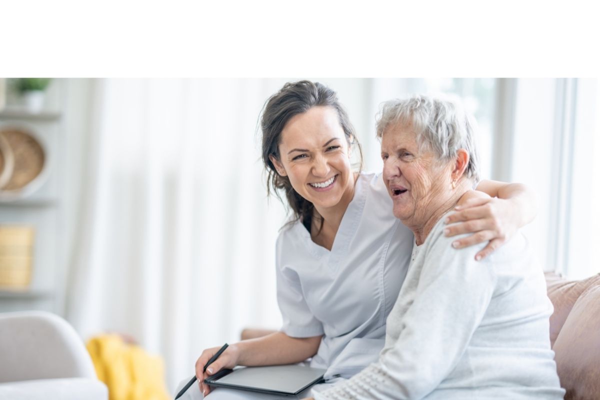 Support worker with a clipboard smiling and hugging an older woman on a couch